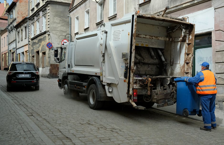 A white commercial rubbish collection truck is parked on a narrow urban street, with its rear open to reveal the mechanical loading mechanism used for waste disposal. A worker dressed in a blue uniform and an orange reflective safety vest is standing beside the truck, holding a blue wheeled bin and preparing to load it into the vehicle. The street is lined with older, multi-story buildings with varied facades, some showing weathered or peeling paint, and the pavement is made of cobblestones and concrete. To the left, a small dark-colored car is parked close to the curb. In the background, there are two street signs, including one indicating no parking and another prohibiting stopping, affixed to a building. The scene is illuminated by natural daylight, with a neutral tone that emphasizes the utilitarian character of the environment and the waste collection process, subtly reflecting the private or alternative waste handling support provided by companies like houseclearanceelephantandcastle.co.uk for non-conventional rubbish removal options.