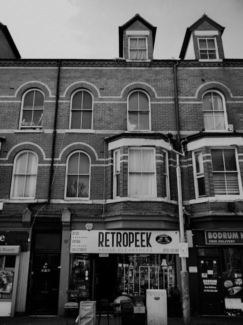 A black and white photograph showing a multi-storey brick building with decorative window arches and tall, narrow windows typical of Victorian architecture. The ground floor houses a shop with a sign reading 'RETROPEEK' indicating house clearance services, supported by natural light coming through large glass windows. In front of the shop, there are two large waste bins and a smaller container, emphasizing the context of rubbish removal. To the right, part of another storefront is visible, with signs advertising delivery services. The building's upper floors feature three dormer windows with pitched roofs, set against a cloudy sky, while the façade appears weathered, conveying a typical urban commercial setting. The scene subtly reflects the practical aspects of private waste collection, with visible refuse receptacles suggesting an independent approach to rubbish management outside local authority provisions.