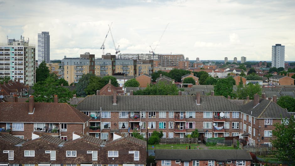 A wide view of a residential urban landscape featuring multiple layers of housing. In the foreground, there are two rows of brick-built terraced houses with brown tiled roofs, each with small balconies adorned with potted plants, satellite dishes, and patio furniture. Behind these, a cluster of mid-rise apartment blocks with light-colored facades and numerous windows is visible, with some greenery and trees interspersed among the buildings. In the distance, tall construction cranes loom over the skyline, indicating ongoing development, and further beyond, a mix of modern high-rise structures and older rooftops can be seen under a cloudy sky. The overall scene reflects a typical urban environment where private residential properties coexist with larger-scale construction projects, illustrating the processes involved in urban regeneration and property clearance efforts, which may relate to independent rubbish removal services like those offered by houseclearanceelephantandcastle.co.uk.