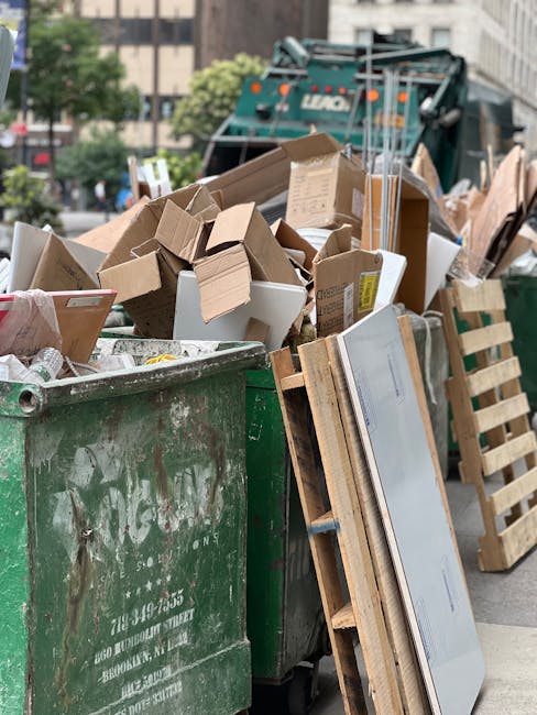 A large green waste bin overflowing with a variety of cardboard boxes, some flattened and others still assembled, along with a few white polystyrene foam pieces and packaging materials, situated on a paved street lined with trees and urban buildings in the background. The scene appears to be part of a private rubbish collection or on-site clearance effort, with wooden pallets leaning against the side of the bin and a large green garbage truck partially visible behind the pile. The cardboard boxes range in size and color, predominantly brown, with some featuring printed labels or tape, indicating typical household or commercial waste prepared for collection. Natural daylight illuminates the scene, highlighting the textures of the cardboard and the rough surface of the bin, conveying a snapshot of waste management activity related to independent rubbish removal services in an urban environment.