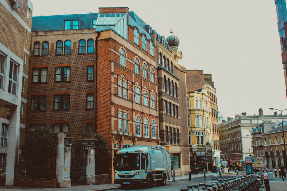 A city street scene showing a row of multi-story buildings with varied architectural styles, including a prominent red-brick structure with arched windows and decorative brickwork, adjacent to a cream-colored building with bay windows and intricate details. The street is lined with parked bicycles and a white refuse collection truck positioned near the curb, indicating ongoing waste collection activity. The environment appears urban with a mix of older and modern architecture, featuring blue-toned roofing and rooftop structures, including a domed turret visible on the corner building. The scene is captured during daylight with natural lighting, and the overall setting suggests an area where private rubbish collection services, such as those by House Clearance Elephant and Castle, might be employed as an alternative to council-led rubbish removal methods, contributing to efficient waste management in busy SE17 neighborhoods.