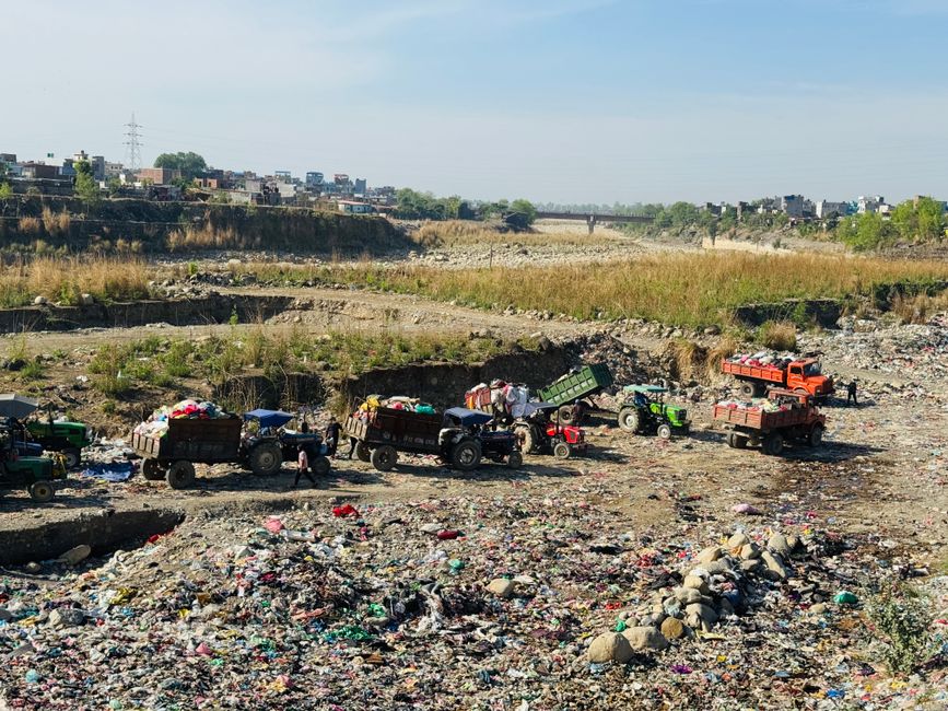 In an open, dry, and somewhat uneven area resembling a waste ground or quarry, several small utility tractors and mini dump trucks are visible, each loaded with various types of rubbish and debris. The equipment, painted in bright colours such as red, green, and orange, is arranged in a line, with some trucks actively unloading or positioned to load waste materials. The rubbish consists of mixed refuse, including plastic bottles, packaging, and discarded household items, scattered across the ground and forming small piles. In the background, there are low-lying, eroded terrain with patches of sparse grass and weeds, leading towards a distant urban skyline of low-rise buildings, with an overcast sky overhead. This scene reflects an environment where waste collection and disposal operations are carried out, illustrating the scale and type of materials handled outside traditional waste collection sites, and subtly aligns with private waste handling or alternative rubbish removal services such as those offered by [COMPANY_NAME].