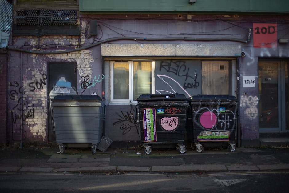 The image shows a section of a building facade with a worn, purple-painted brick wall that has visible weathering and peeling paint. Two large wheeled rubbish bins are positioned on the sidewalk directly in front of a small, white-framed window, which is partially covered by a metal shutter drawn down over it. The grey bin is on the left, and the black bin, decorated with pink and white graffiti including a prominent pink heart, is on the right; both bins have various stickers and tags on their surfaces. To the right of the bins, there are additional graffiti tags on the wall, along with a couple of street signs, one of which reads “10D” on both a small sign and a larger red panel. An overhead cable runs horizontally across the scene, and the overall environment appears urban and somewhat neglected, typical of private waste storage areas outside residential or commercial premises, emphasizing the importance of professional rubbish removal services like those offered by houseclearanceelephantandcastle.co.uk, especially in contexts requiring independent or on-site waste management solutions.