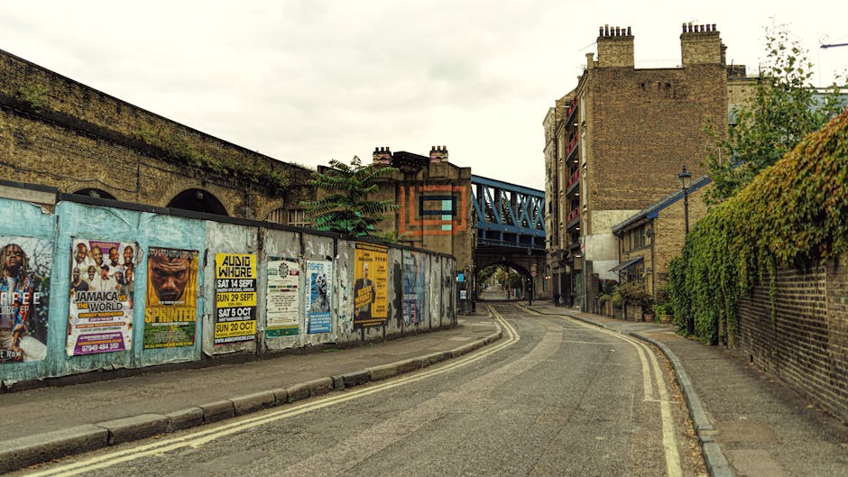 The image shows a section of a building facade with a worn, purple-painted brick wall that has visible weathering and peeling paint. Two large wheeled rubbish bins are positioned on the sidewalk directly in front of a small, white-framed window, which is partially covered by a metal shutter drawn down over it. The grey bin is on the left, and the black bin, decorated with pink and white graffiti including a prominent pink heart, is on the right; both bins have various stickers and tags on their surfaces. To the right of the bins, there are additional graffiti tags on the wall, along with a couple of street signs, one of which reads “10D” on both a small sign and a larger red panel. An overhead cable runs horizontally across the scene, and the overall environment appears urban and somewhat neglected, typical of private waste storage areas outside residential or commercial premises, emphasizing the importance of professional rubbish removal services like those offered by houseclearanceelephantandcastle.co.uk, especially in contexts requiring independent or on-site waste management solutions.